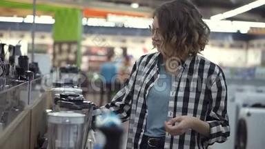 在电器商店里，一位穿着格子衬衫的黑发卷发女士通过观看和拿着来选择搅拌机购物